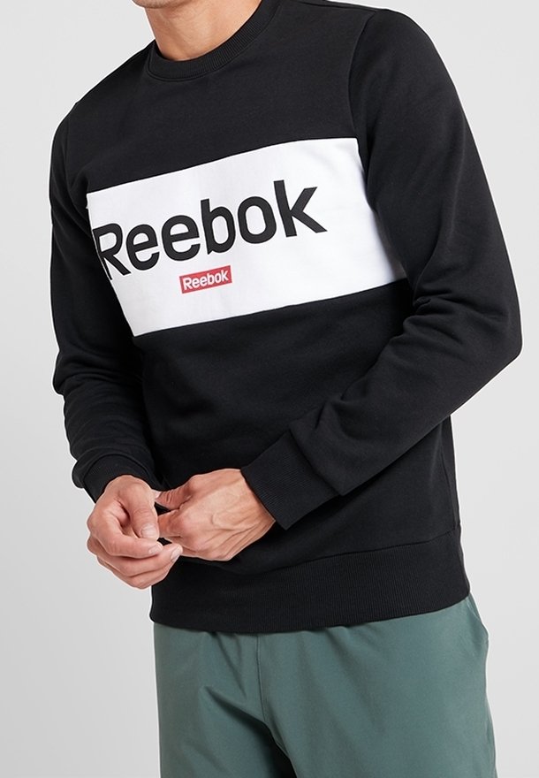 Homme portant un sweat-shirt Reebok noir et blanc avec un logo sur la poitrine et un pantalon vert, ajustant ses manches.