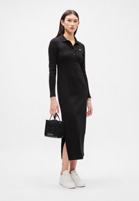 Μαύρο φόρεμα με μακριά μανίκια και κολάρο, με τρία κουμπιά, που διαθέτει πλάγια σχισμή και συνδυάζεται με μία μικρή μαύρη τσάντα και λευκά αθλητικά παπούτσια.