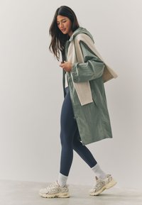 Lightweight, green jacket with cream accents, paired with navy leggings and beige athletic shoes. The jacket has a hood and a relaxed fit.