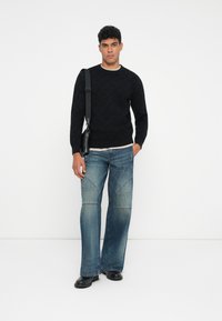 Mann in schwarzem Pullover, blauen Schlagjeans mit Knieabnähten, schwarzen Schuhen und einer schwarzen Umhängetasche vor einem weißen Hintergrund.