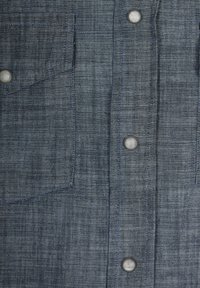 Camicia di lino blu con tessuto testurizzato, dotata di taschino sul petto e bottoni a pressione argentati lungo la parte anteriore.