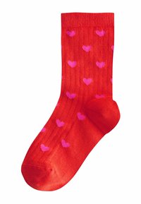 Sarkana zeķe ar nelielu rozā sirdīšu rakstu, kas vienmērīgi sadalīts pa visu audumu, ar ribotu aproci augšpusē.