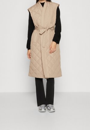 Femme portant une longue veste sans manches beige matelassée avec une ceinture, un haut noir à manches longues, un pantalon noir et des baskets argentées, debout contre un mur blanc.