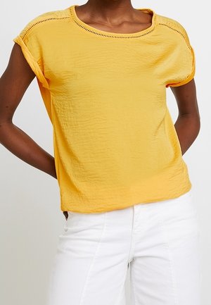 Woman wearing a short-sleeve yellow textured top with rolled cuffs and white high-waisted pants, hands behind her back.