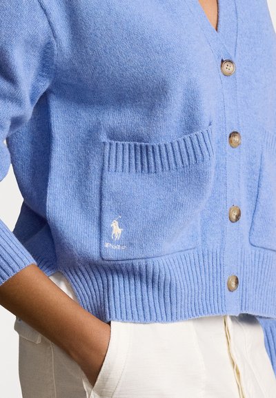 Gaiši zils kardigans, kas izgatavots no mīksta adījuma materiāla. Tam ir V-veida kakls, ribota piedurkne, divas priekšējās kabatas un logo uz kreisās kabatas.