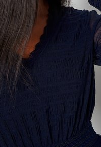 Robe bleu marine en tissu texturé, dotée d'un col en forme de vagues et de détails froncés. Les manches courtes mettent en valeur le design.