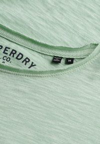 Primer plano del escote de una camiseta Superdry verde claro que muestra la textura de la tela, etiqueta interna etiquetada como "Hecho en India" y etiqueta de tamaño M.