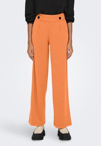 Oranje wijde broeken met een hoge taille, voorzien van twee zwarte decoratieve knopen en een gladde, gestructureerde stof. Gepaard met zwarte schoenen.