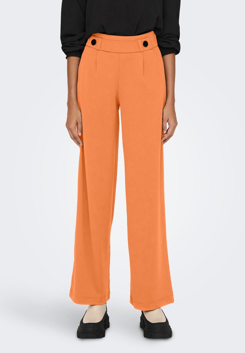 Oranje wijde broeken met een hoge taille, voorzien van twee zwarte decoratieve knopen en een gladde, gestructureerde stof. Gepaard met zwarte schoenen.