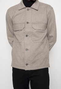 Beige knoopjasje met twee voorzakken, gedragen over een zwarte coltrui en gecombineerd met zwarte broek.