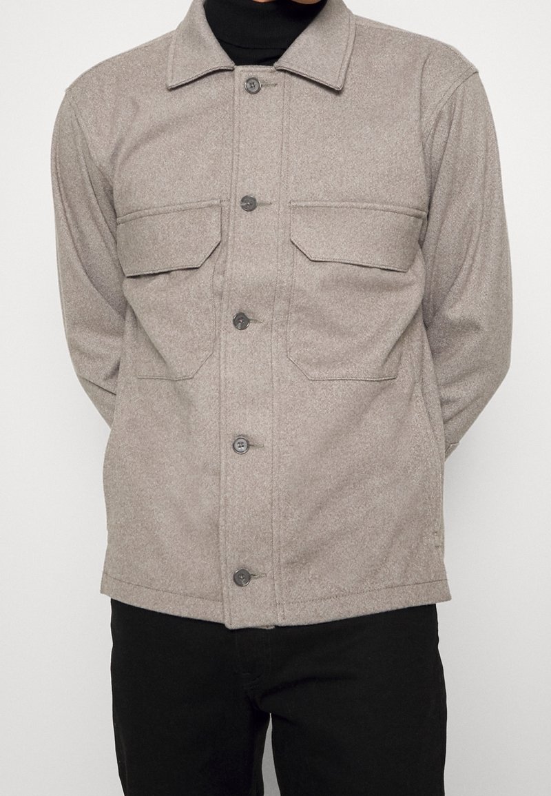 Beige knoopjasje met twee voorzakken, gedragen over een zwarte coltrui en gecombineerd met zwarte broek.