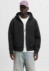 Sort zip-up hoodie i blødt materiale med en frontlomme, ribbet kant og en hvid grafisk t-shirt under. Lyseblå jeans og en lyserød kasket.