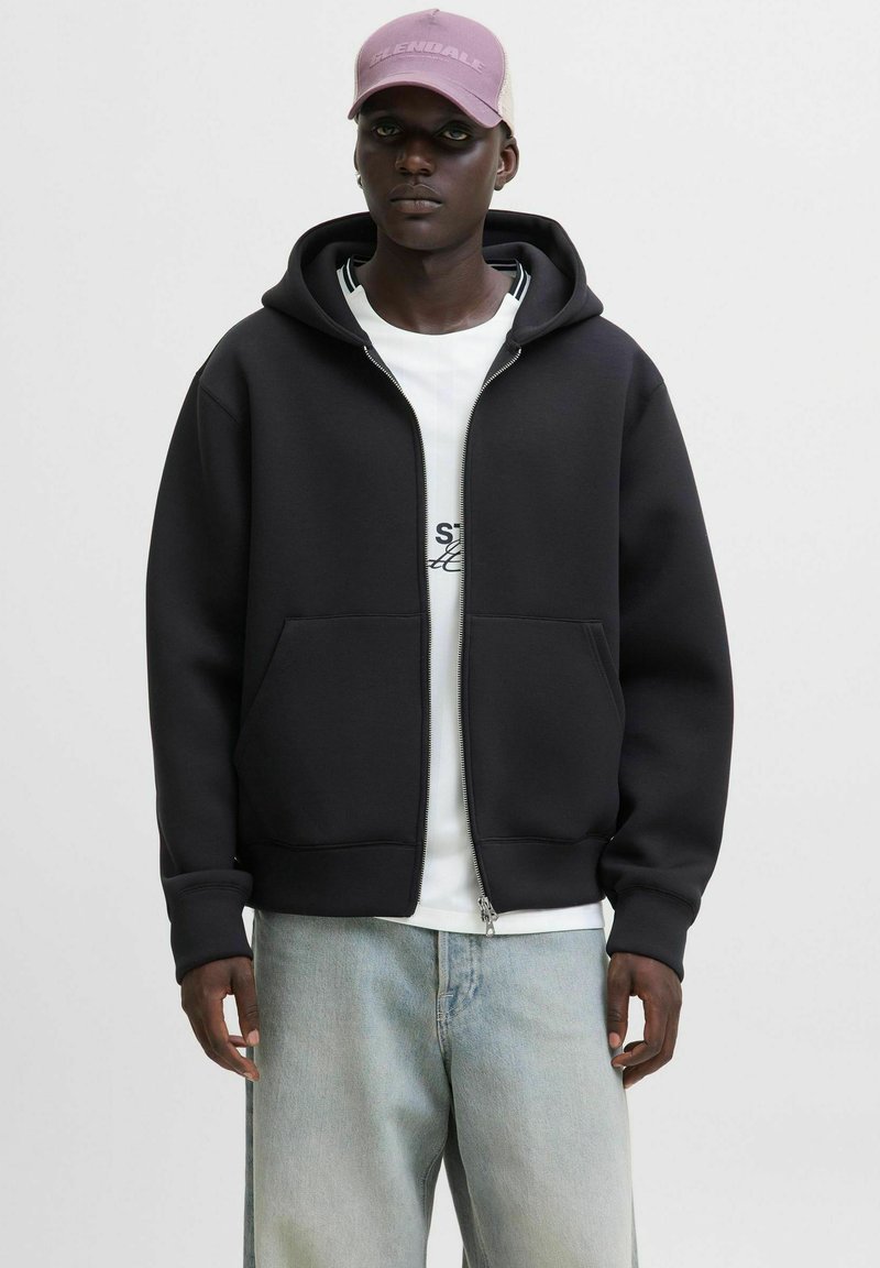 Sort zip-up hoodie i blødt materiale med en frontlomme, ribbet kant og en hvid grafisk t-shirt under. Lyseblå jeans og en lyserød kasket.