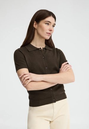 Mujer con cabello largo y castaño, vistiendo una camisa de manga corta marrón oscuro abotonada y pantalones claros, de pie con los brazos cruzados mirando hacia la derecha.