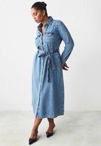 Denim-paita mekko pitkillä hihoilla, kauluksella, etutaskuilla, vyötärölle sidottuna ja polvien keskikohdan pituudella vaaleansinisessä pesussa. Mustat kärkitossut käytössä.