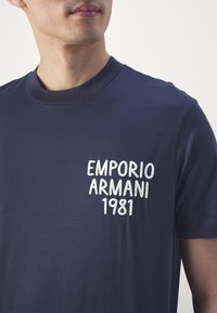 Námořnická modrá bavlněná trička s krátkým rukávem, na levé hrudi s bílým vyšitým textem "EMPORIO ARMANI 1981".