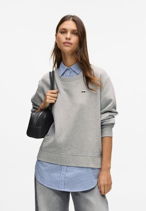 Młoda kobieta w warstwowym szarym swetrze na niebieskiej koszuli w kratkę, trzymająca czarną torebkę na ramieniu, na tle białego tła.