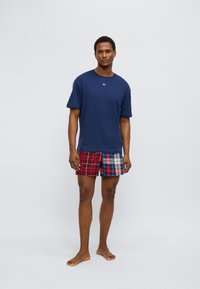 Μπλε ναυτικό βαμβακερό μπλουζάκι με κοντά μανίκια και μικρό λογότυπο· κόκκινο και μπλε καρό μαγιό σορτς με αντιθετικά σχέδια· γυμνά πόδια.