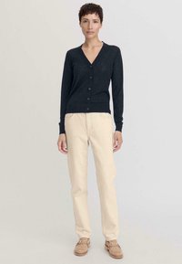 Gilet en laine navy avec un col v et six boutons, associé à un jean couleur crème. Modèle portant des mocassins beige, se tenant contre un fond neutre.