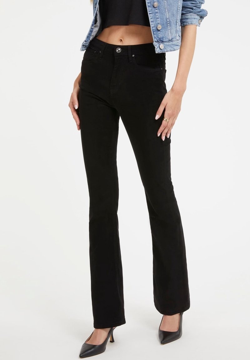 Femme portant un jean noir taille haute à jambes droites, un top noir court, une veste en denim bleu clair et des talons noirs à bout pointu sur fond blanc.