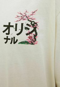 Svetlo bež majica s grafičnim motivom veće cveta u roza in zeleni barvi ter črnim japonskim besedilom spodaj.