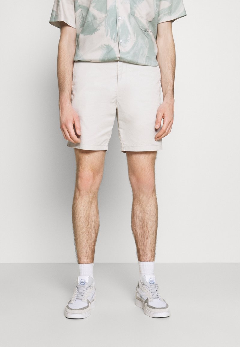 Shorts beige clair en mélange de coton, avec une taille plate, des poches latérales et une coupe décontractée, associés à des baskets blanches et des chaussettes.