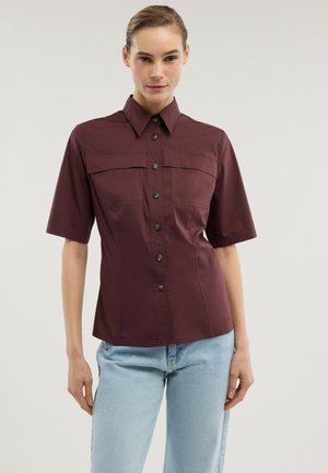 Kvinde iført en knappet, kortærmet, mørk burgunderfarvet skjorte med brystlommer og lyseblå jeans, stående mod en hvid baggrund.