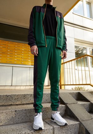 Person iført grøn og sort Nike træningsdragt med hvide sneakers, der står på betontrapper udenfor en bygning med gule postkasser.