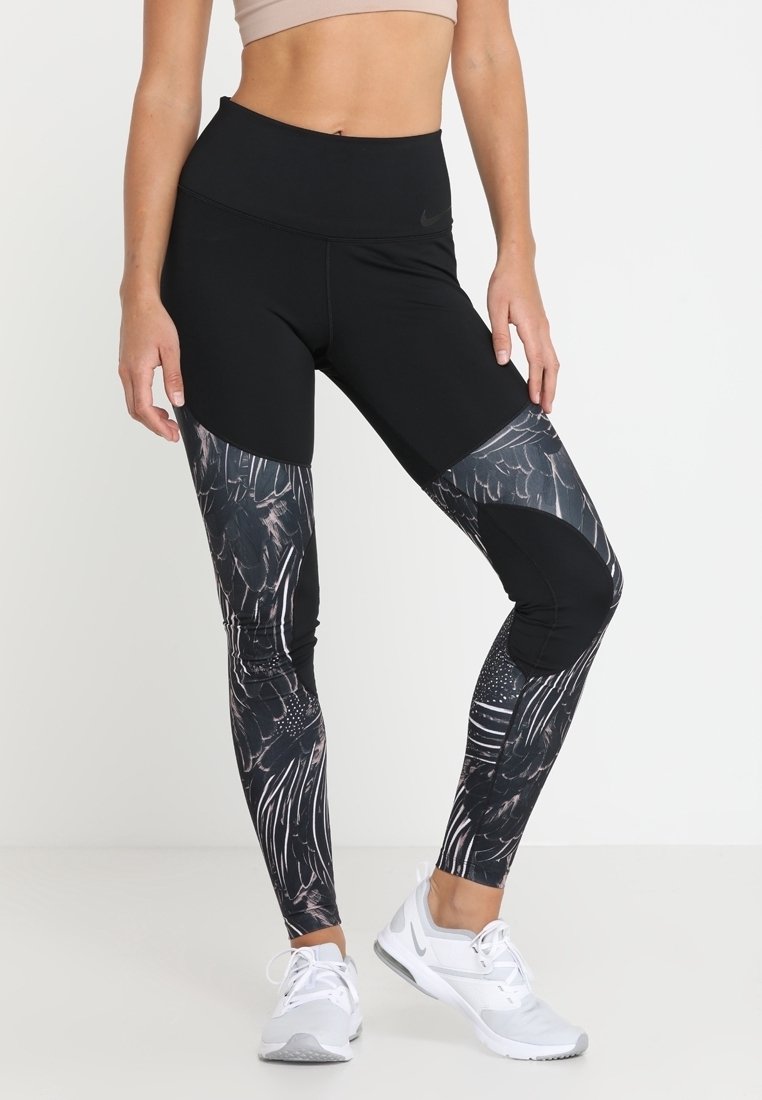 Donna che indossa leggings neri a vita alta con motivi astratti a forma di piuma sulle gambe e scarpe da ginnastica bianche, in piedi contro uno sfondo bianco.