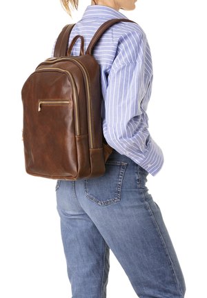Persoon draagt een blauw gestreept shirt en spijkerbroek met een bruine leren rugzak met meerdere met ritsen afsluitbare vakken.