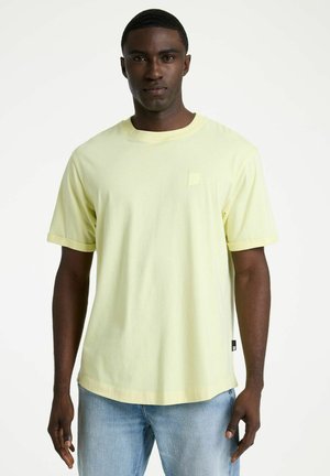 Muž v svetložltom tričku s krátkym rukávom a svetlo modrých džínsoch stojí pred jednoliatym bielym pozadím.