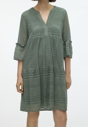 Spitzenkleid in gedämpftem Grün mit V-Ausschnitt, 3/4-Ärmeln und kunstvollen Mustern sowie einem gestuften Saum. Leichter, strukturierter Stoff.