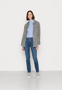 Casaco xadrez em preto e branco sobre um suéter de gola alta azul claro, combinado com jeans azuis justos e ténis cinzentos. Fundo branco liso.