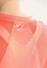 Nike-Sportshirt in hellem Korallenrot mit Mesh-Struktur, versehen mit einem dezenten silbernen Logo in der Nähe des Halsausschnitts und kontrastierenden Nahtdetails.