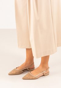 Bailarinas beige con tiras en el talón, punta afilada, detalles de malla y un pequeño tacón. Presentan una correa de tobillo en tono marrón con un acento de hebilla.