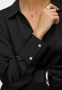 Crna košulja s glatkom teksturom, koja ima ovratnik i manžete s dugmadima bijele boje. Srebrni prsten i ogrlica upotpunjuju izgled.
