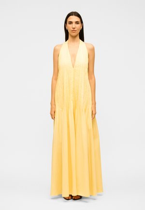 Γυναίκα με ίσια σκουρόχρωμα μαλλιά, που φοράει κίτρινο μίντι φόρεμα με πιέτες, χωρίς μανίκια και μαύρα σανδάλια, στέκεται μπροστά από απλό φόντο.