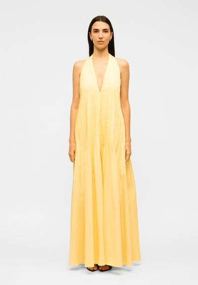 Femme aux cheveux bruns et raides, portant une robe maxi plissée jaune sans manches avec un col halter et des sandales noires, debout devant un fond uni.