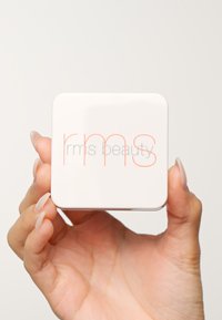 Vierkante witte compact met afgeronde hoeken, met "rms beauty" in grijze en oranje tekst. Vervaardigd in een hand met lichtgekleurde nagels.
