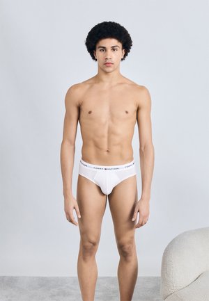 Baltas kokvilnas biksītes ar melnu jostu, kurā ir „TOMMY HILFIGER” logo; modelis stāv gaiši pelēka fona priekšā.