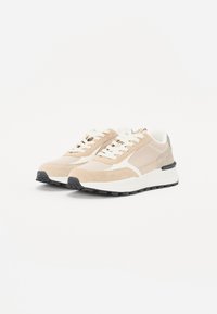 Beige semskinnsneakers med mesh-detaler, hvite lisser, en polstret såle og en svart gummisåle. Har et glatt design og teksturerte detaljer.