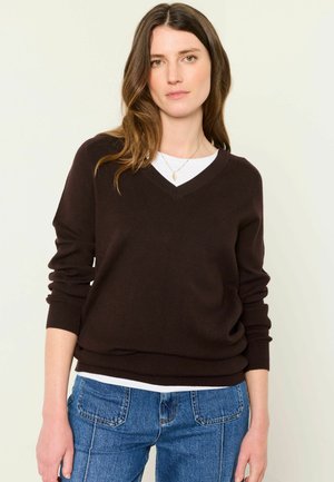 Giovane donna con lunghi capelli castani che indossa un maglione a V marrone scuro sopra una canottiera bianca e jeans blu su uno sfondo chiaro e semplice.