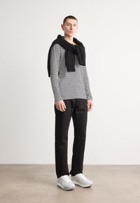 Stripet langermet skjorte i svart og hvitt, svart genser knyttet rundt nakken, svarte bukser og lysegrå joggesko. Minimalistisk design.