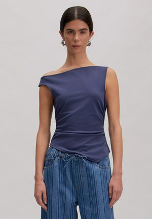 Vrouw met donker haar in een mouwloze asymmetrische marineblauwe top en hooggetailleerde blauw gestreepte spijkerbroek tegen een effen lichte achtergrond.