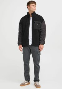 Chaqueta de forro polar negra y gris con un collar alto, un bolsillo en el pecho y una textura suave, combinada con pantalones cargo grises y zapatillas.