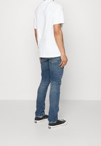 Blå denimjeans med en slim fit, bärs med en enkel vit t-shirt, tillsammans med svarta sneakers. Jeansens har traditionella fickor och sömmar.