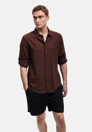 Bruine button-up shirt met lange mouwen opgerold, gemaakt van een gladde stof. Gecombineerd met zwarte shorts. Eenvoudig ontwerp, geen zichtbare patronen of accenten.