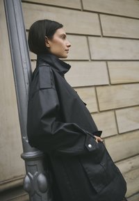 Čierna nepremokavá bunda s vysokým golierom, veľkými vreckami, gombíkovými detailami a voľným strihom, fotografovaná na svetlom pozadí.