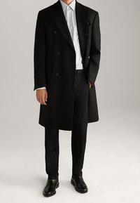 Cappotto nero doppiopetto in lana strutturata, con revers a découpage sopra una camicia a righe bianca e blu, abbinato a pantaloni e scarpe neri.