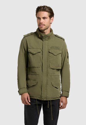 Férfi, aki olíva zöld színű, katonai stílusú dzsekit visel, amelyen több zseb és vállpánt található, fekete nadrággal, szürke háttér előtt áll.